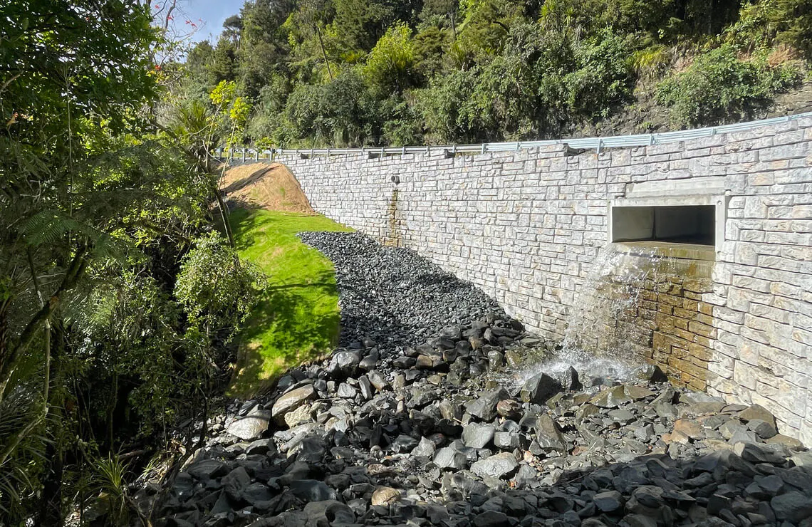 Water flows from a drainage outlet in a Ledgestone-textured Redi-Rock retaining wall beside a forested New Zealand roadway.