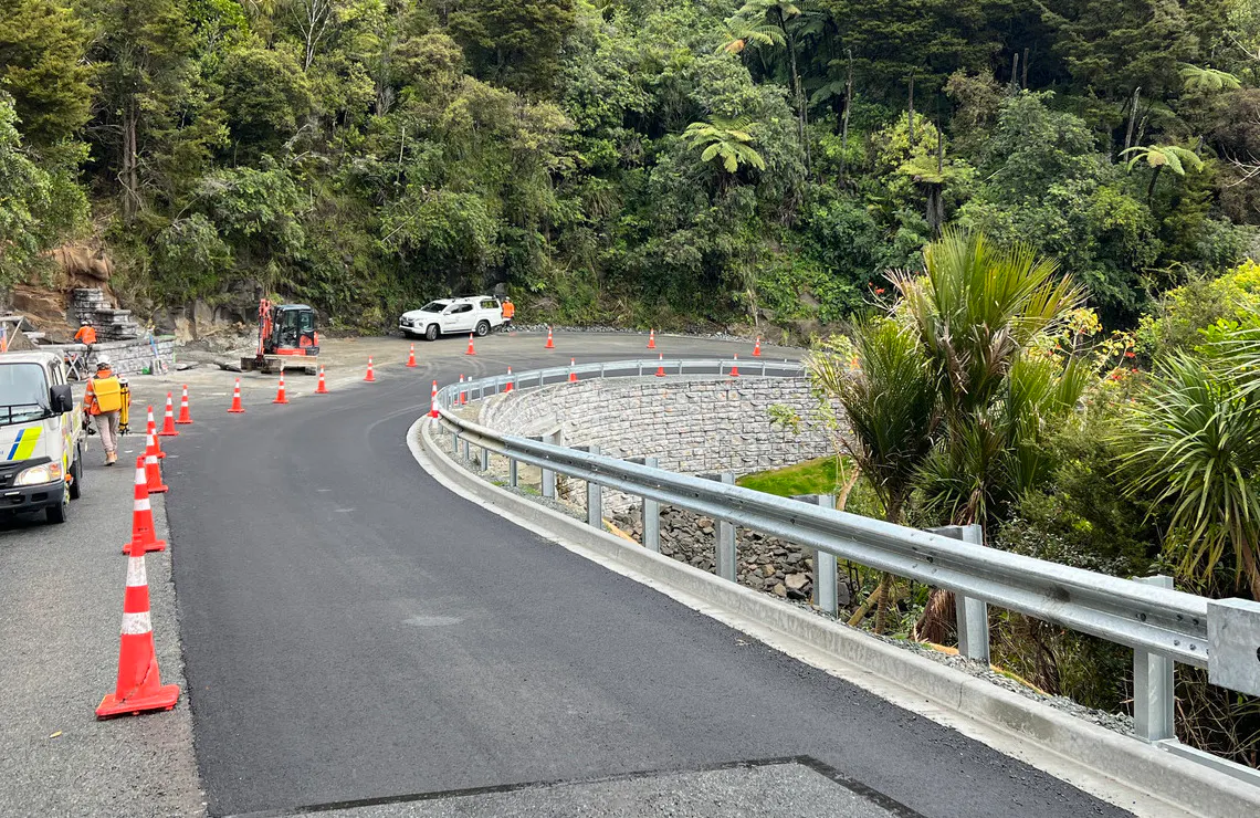 Road crews complete paving beside a curved Redi-Rock hybrid retaining wall on a forested New Zealand roadway.