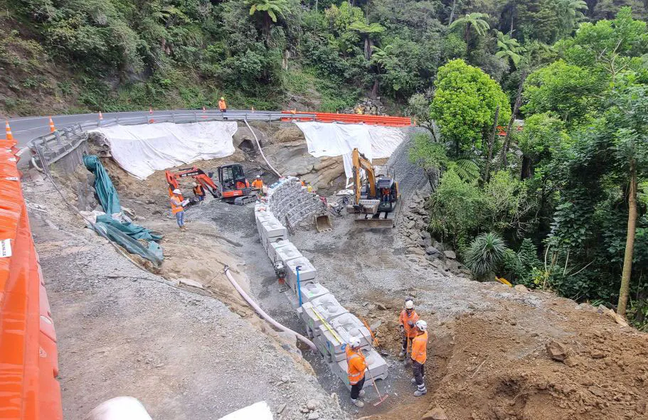 Crews use excavators to install a Redi-Rock hybrid retaining wall beside a forested New Zealand roadway.
