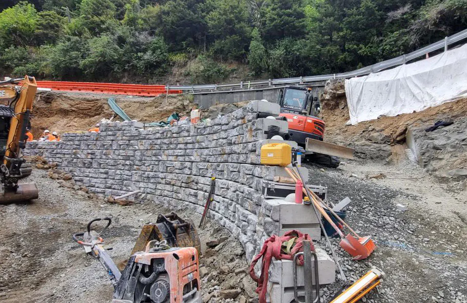 Excavators and crews construct a Ledgestone-textured Redi-Rock hybrid retaining wall below a forested New Zealand roadway.